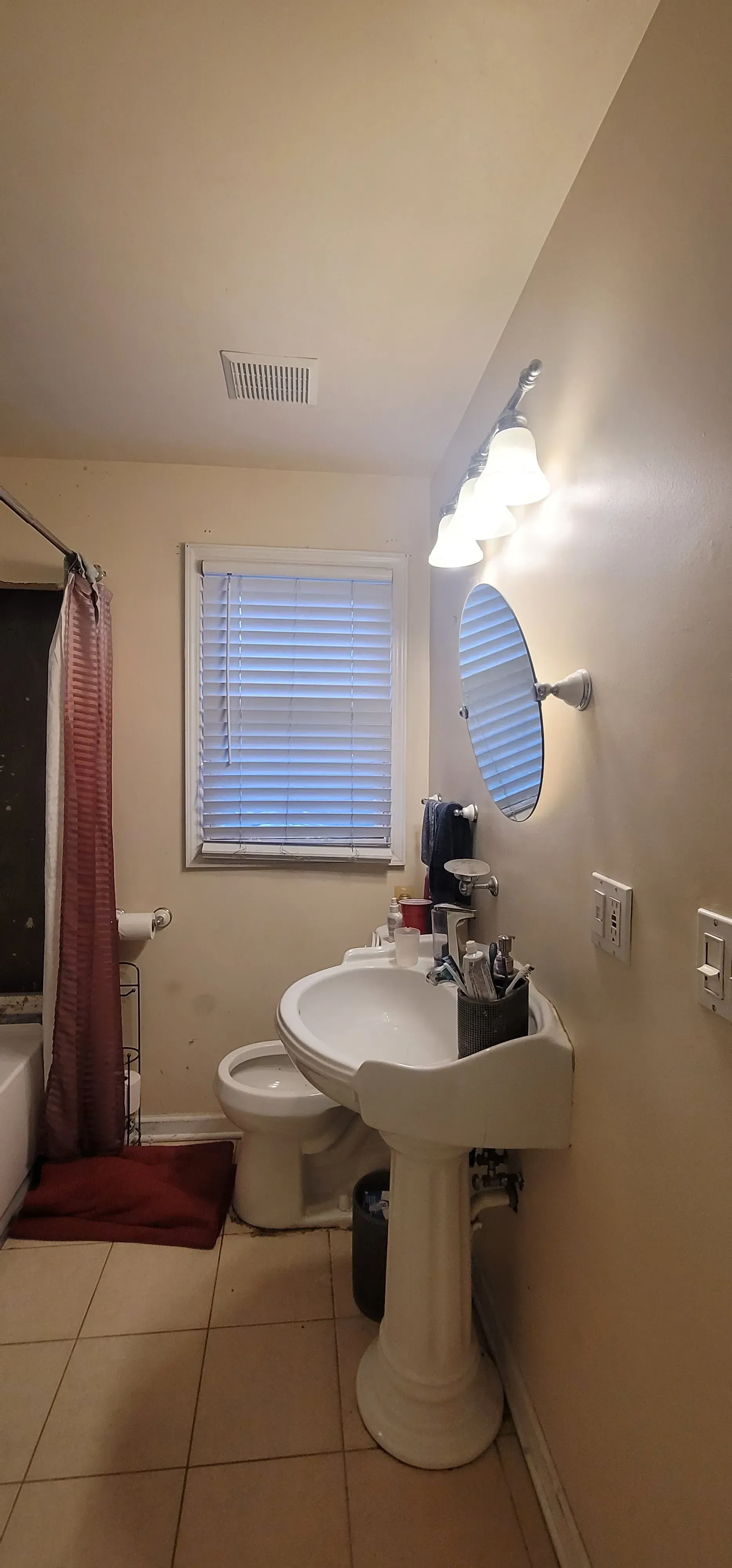 A small, cream-colored bathroom with a pedestal sink, toilet, and window with blinds