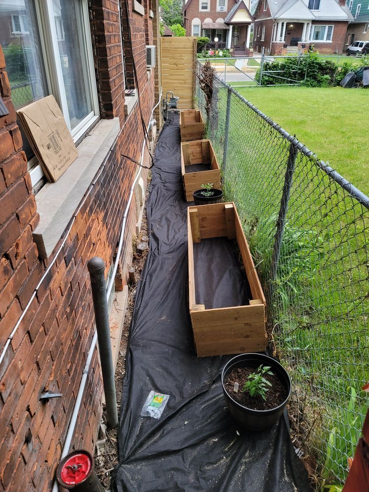 Wooden planters line a narrow space between a brick wall and a chain-link fence