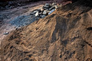Pile of brown soil with gravel, stones, and darker areas in outdoor setting.