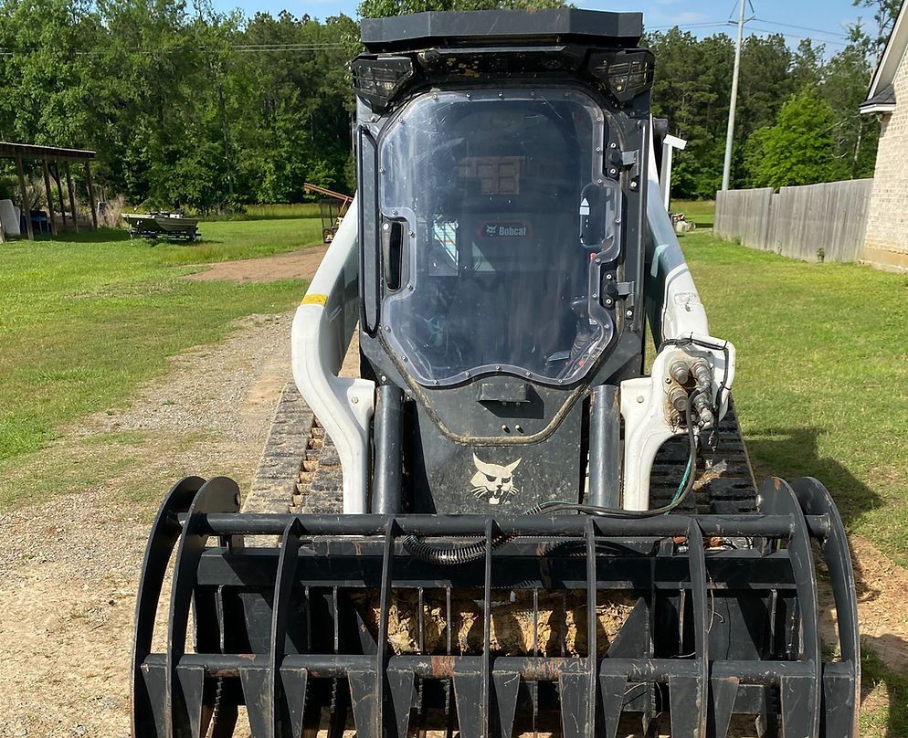 Bobcat skid-steer loader with brush grapple attachment; outdoors on dirt, grass, and trees in background.