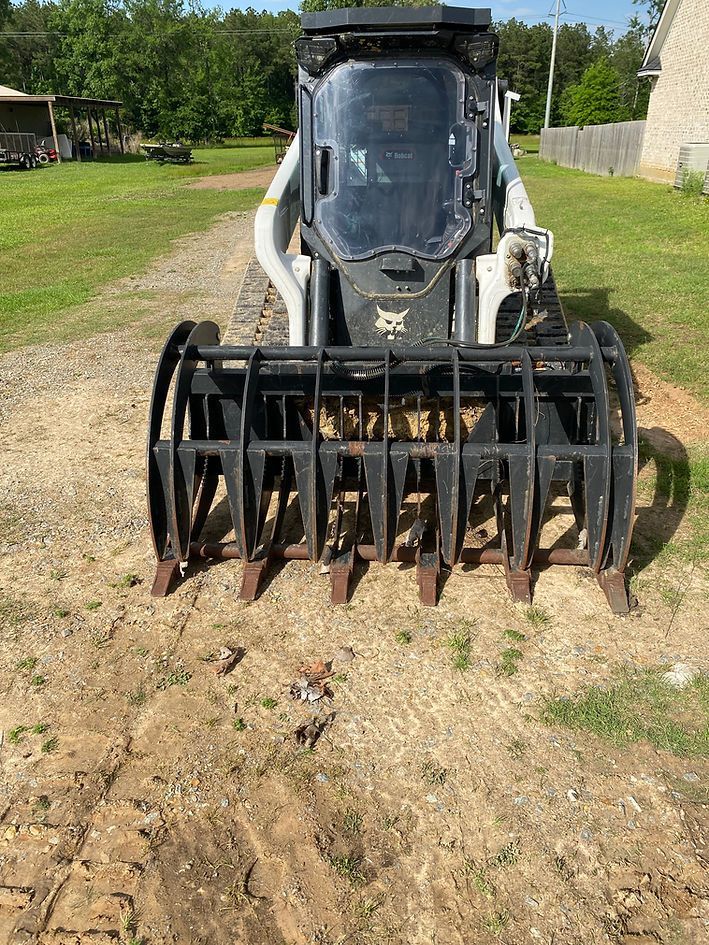 Bobcat skid-steer loader with a grapple attachment, sitting on dirt and grass.
