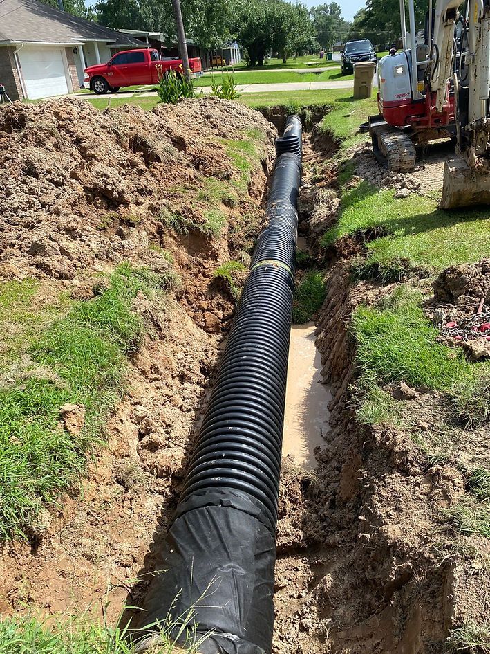Black corrugated drainage pipe in a trench, beside a small excavator and residential homes.