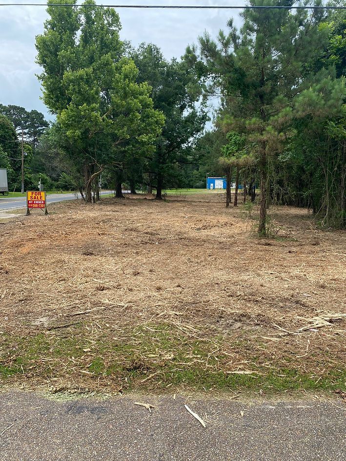 Cleared lot with a For Sale sign, bordered by trees and a roadway.