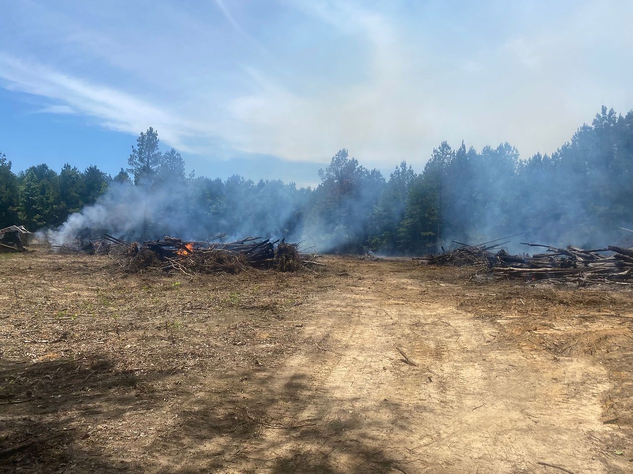 Pile of debris burning in a field, smoke rising toward the blue sky and trees.