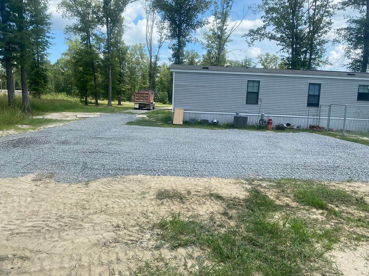 Gravel driveway in front of a gray mobile home. A truck is parked further back on the driveway.