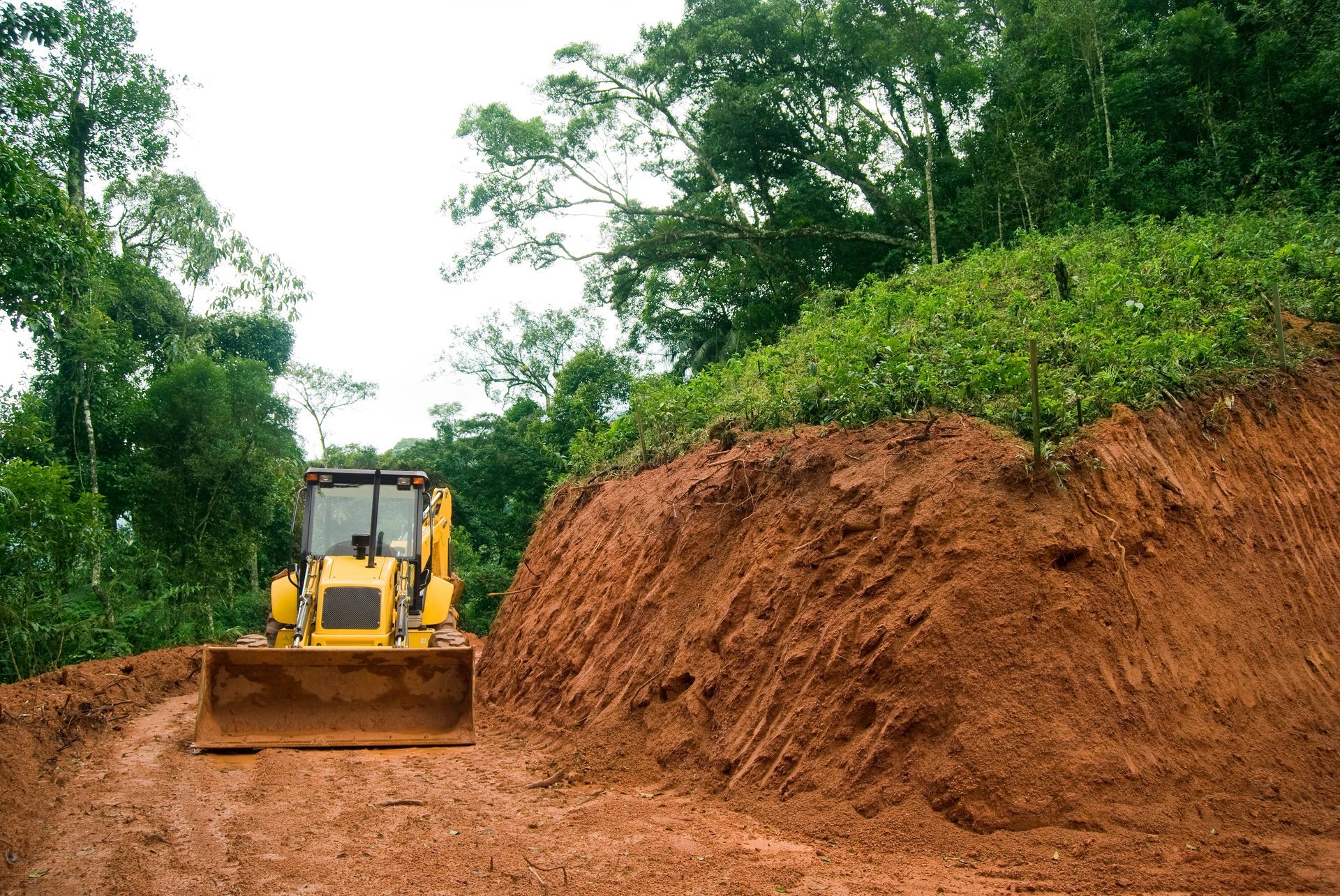 Yellow bulldozer clearing a muddy red earth road through a forested hillside.
