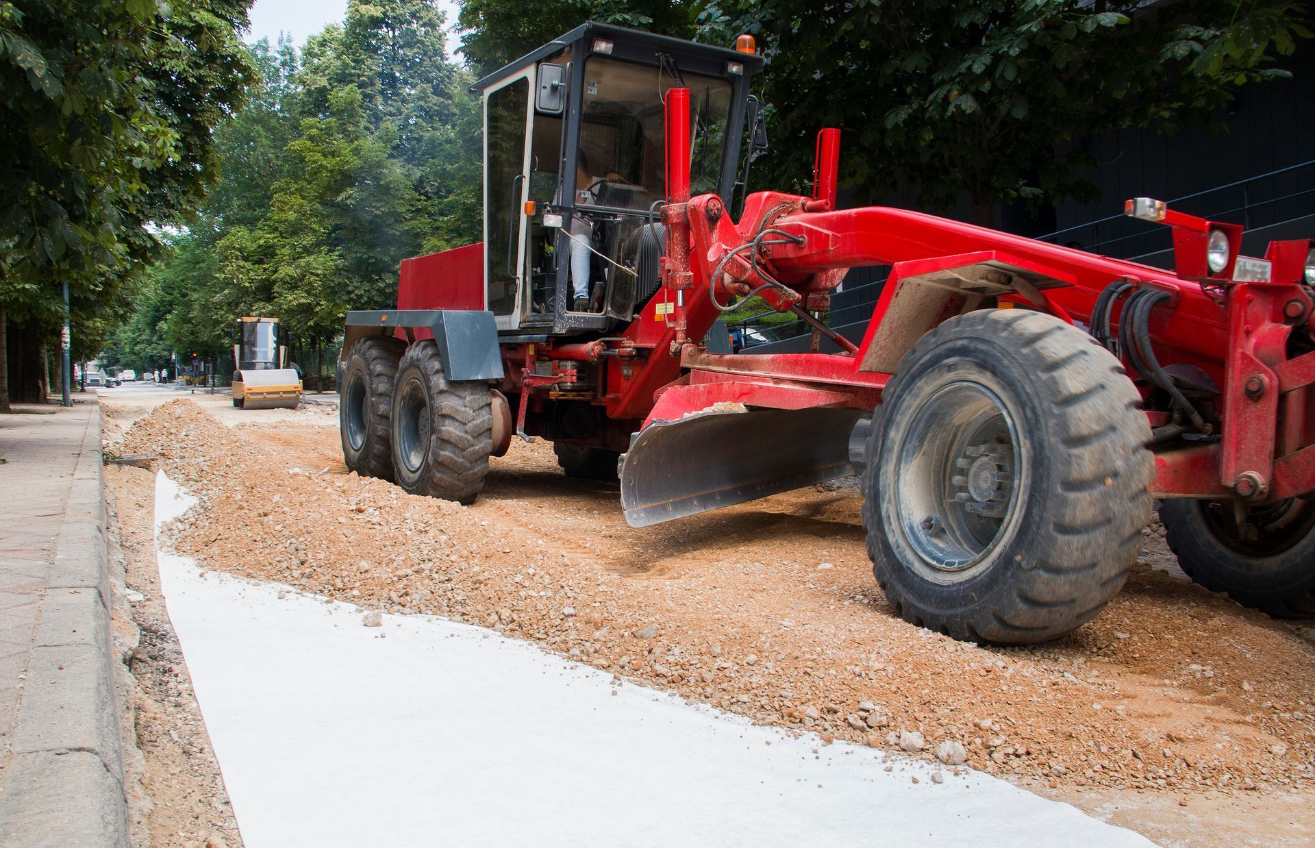 Red road grader smoothing gravel on a city street. Trees and a small roller are in the background.