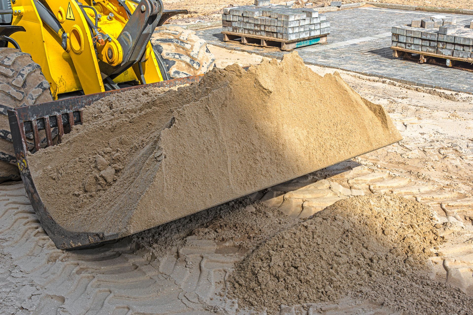 Yellow loader bucket filled with tan sand on a construction site, pavers in the background.