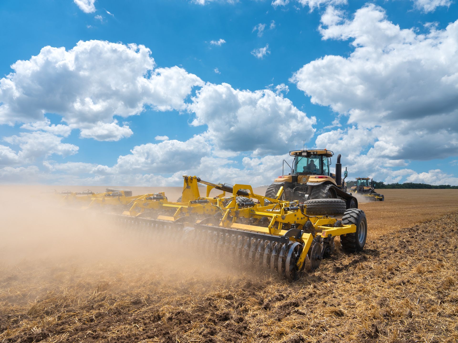 Yellow tractor plowing a field under a blue sky with fluffy clouds, creating dust.