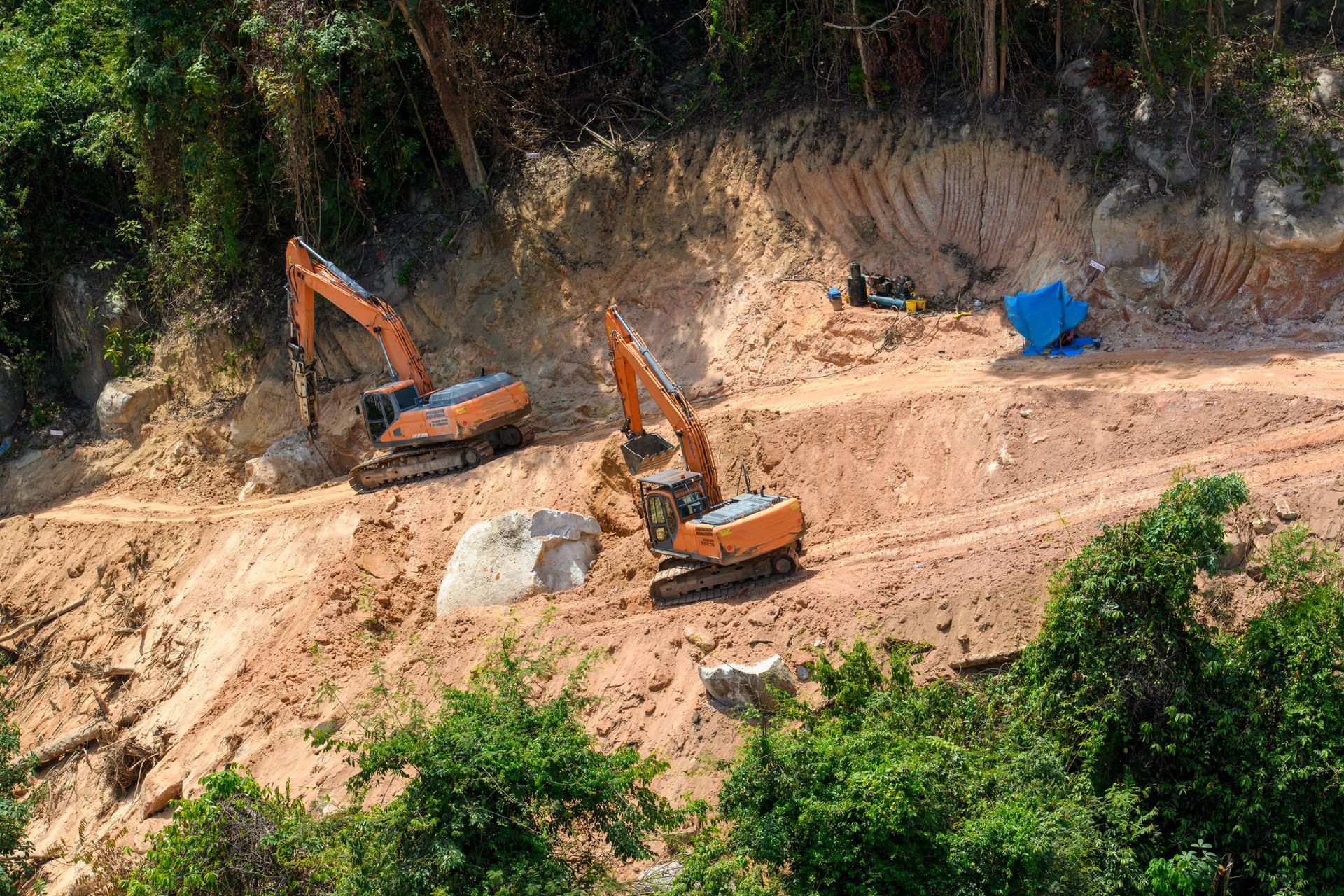 Two orange excavators on a hillside, clearing dirt and rocks for construction, amidst green trees and blue tarp.