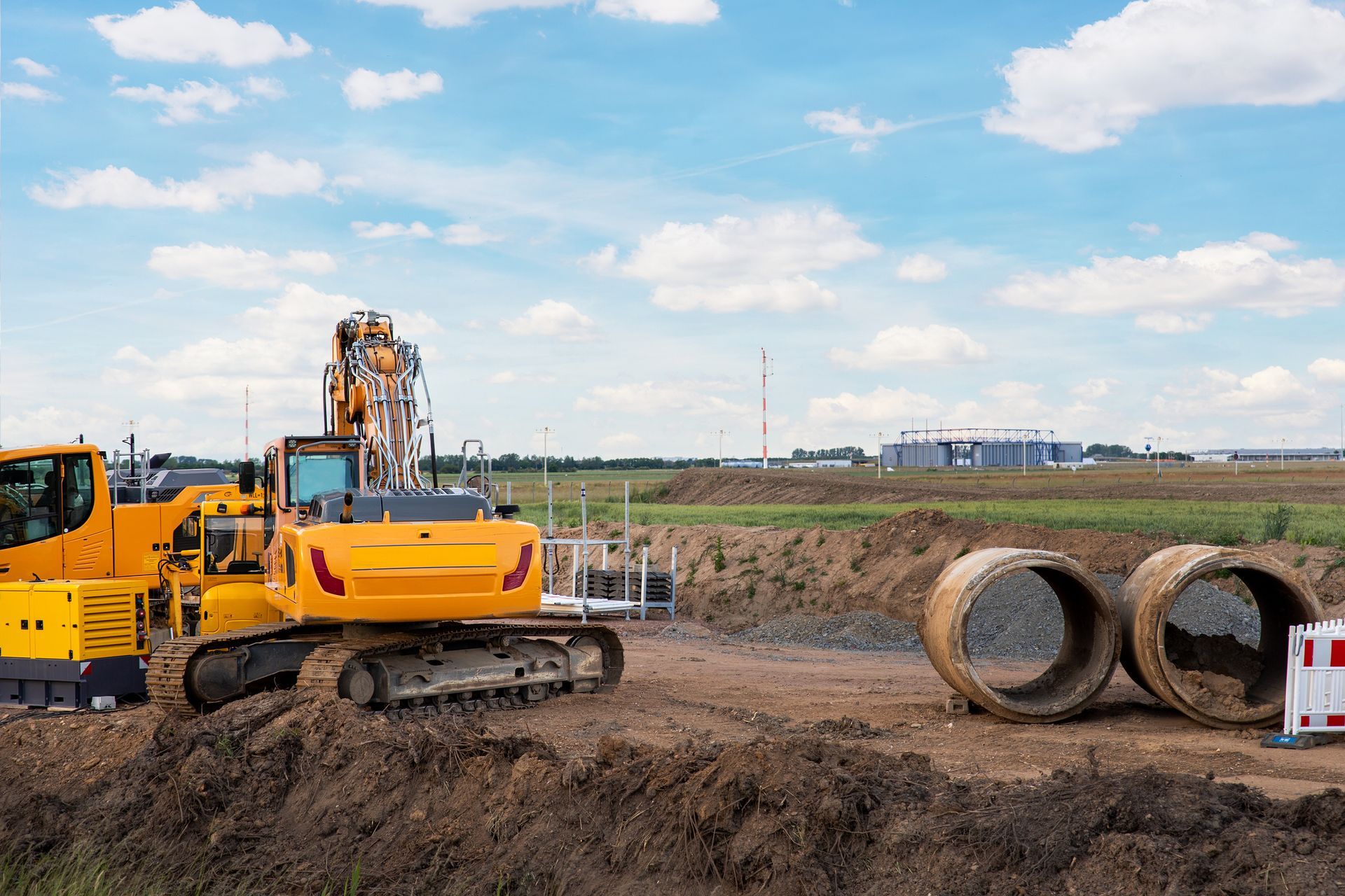 Yellow excavator on a construction site with large concrete pipes, sunny day, blue sky.