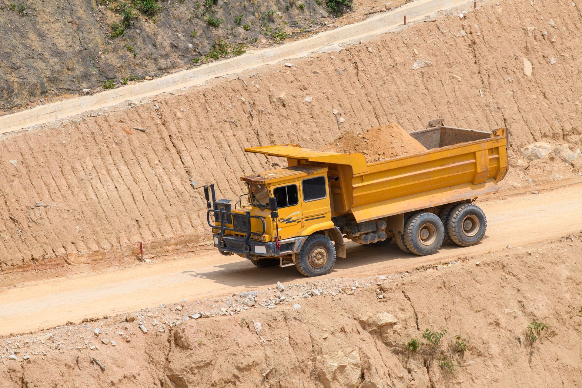 Yellow dump truck carrying dirt on a dirt road cut into a hillside.