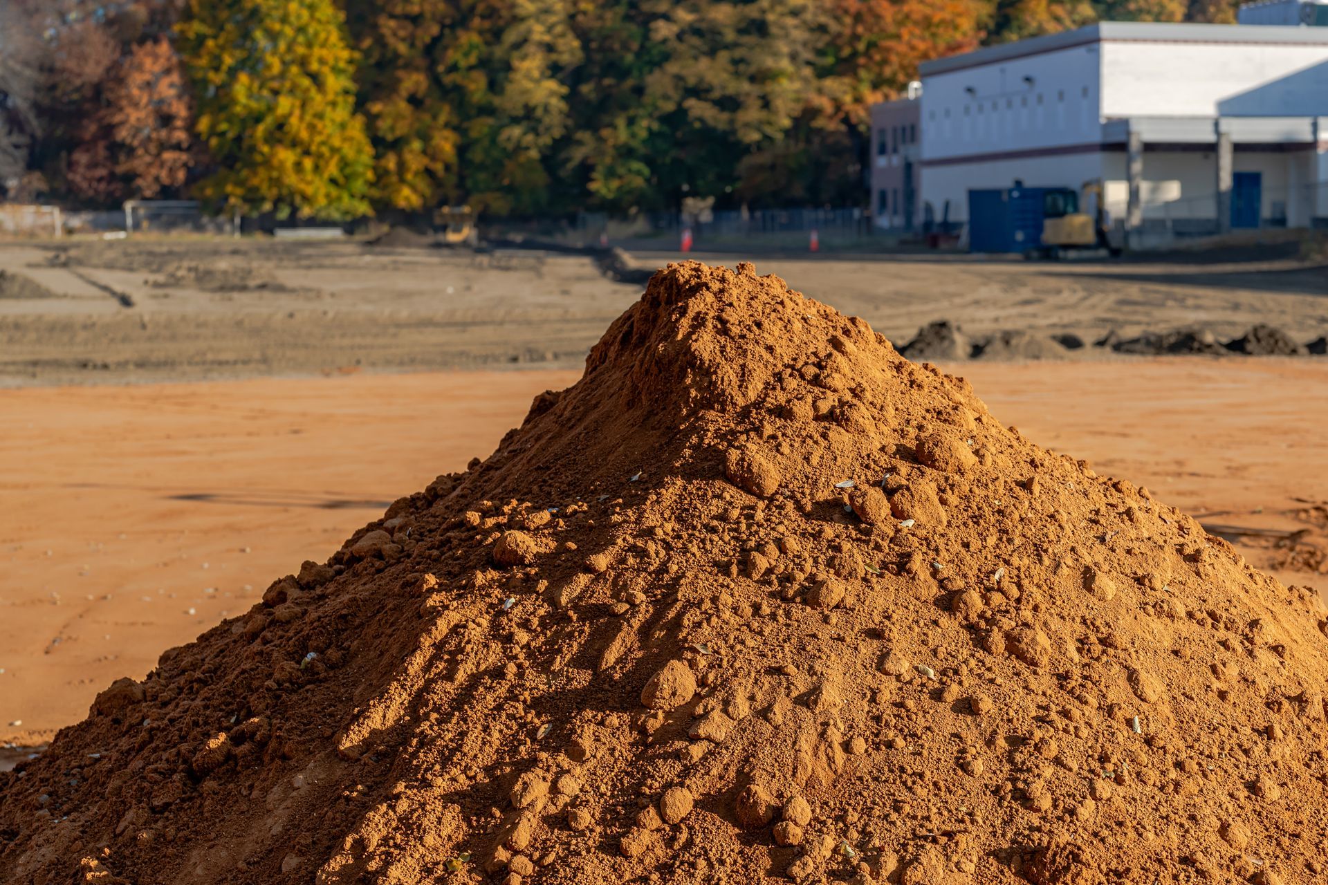 Pile of brown dirt on a construction site; building and trees in the background.