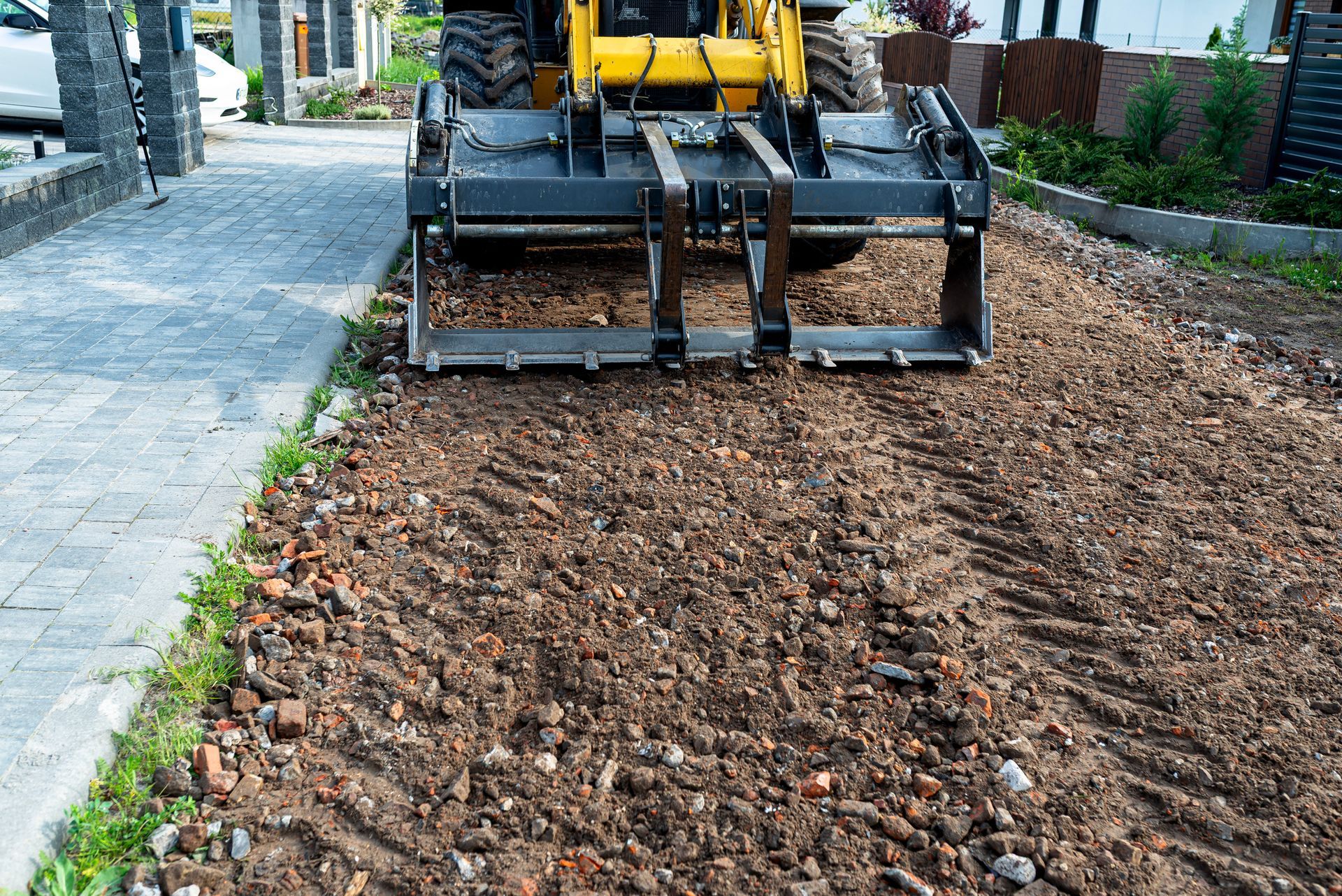 A yellow construction vehicle with a bucket claw is working on a dirt and brick driveway next to a brick sidewalk.
