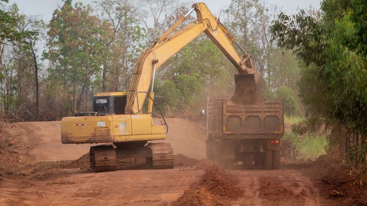 Yellow excavator loading dirt into a dump truck on a dirt road, trees in the background.