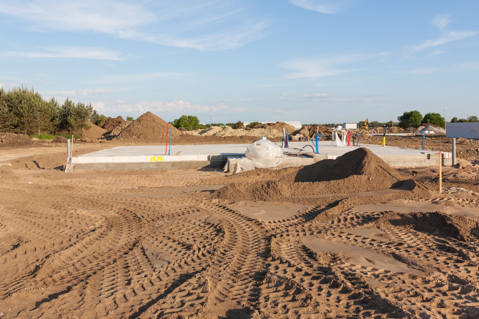 Construction site with a concrete foundation, dirt piles, and tire tracks under a blue sky.