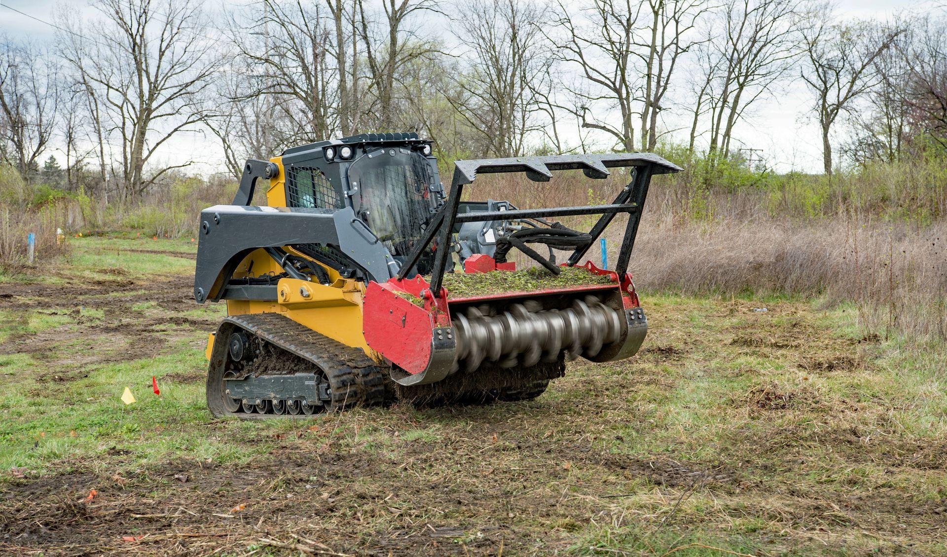 Yellow skid-steer mulching vegetation in a field with bare trees in the background.
