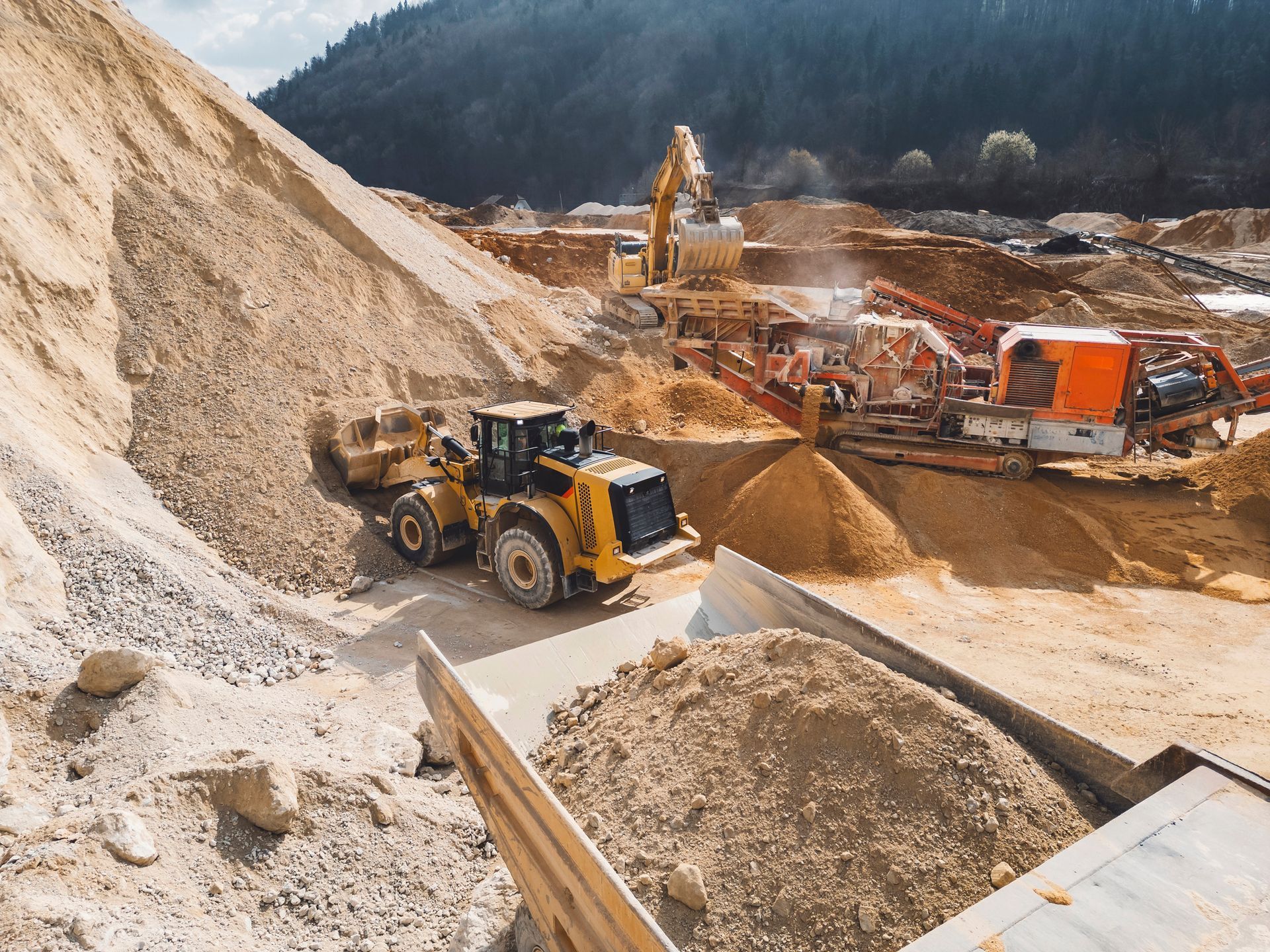A wheel loader at a quarry moves material near a processing machine; hills and trees in the background.