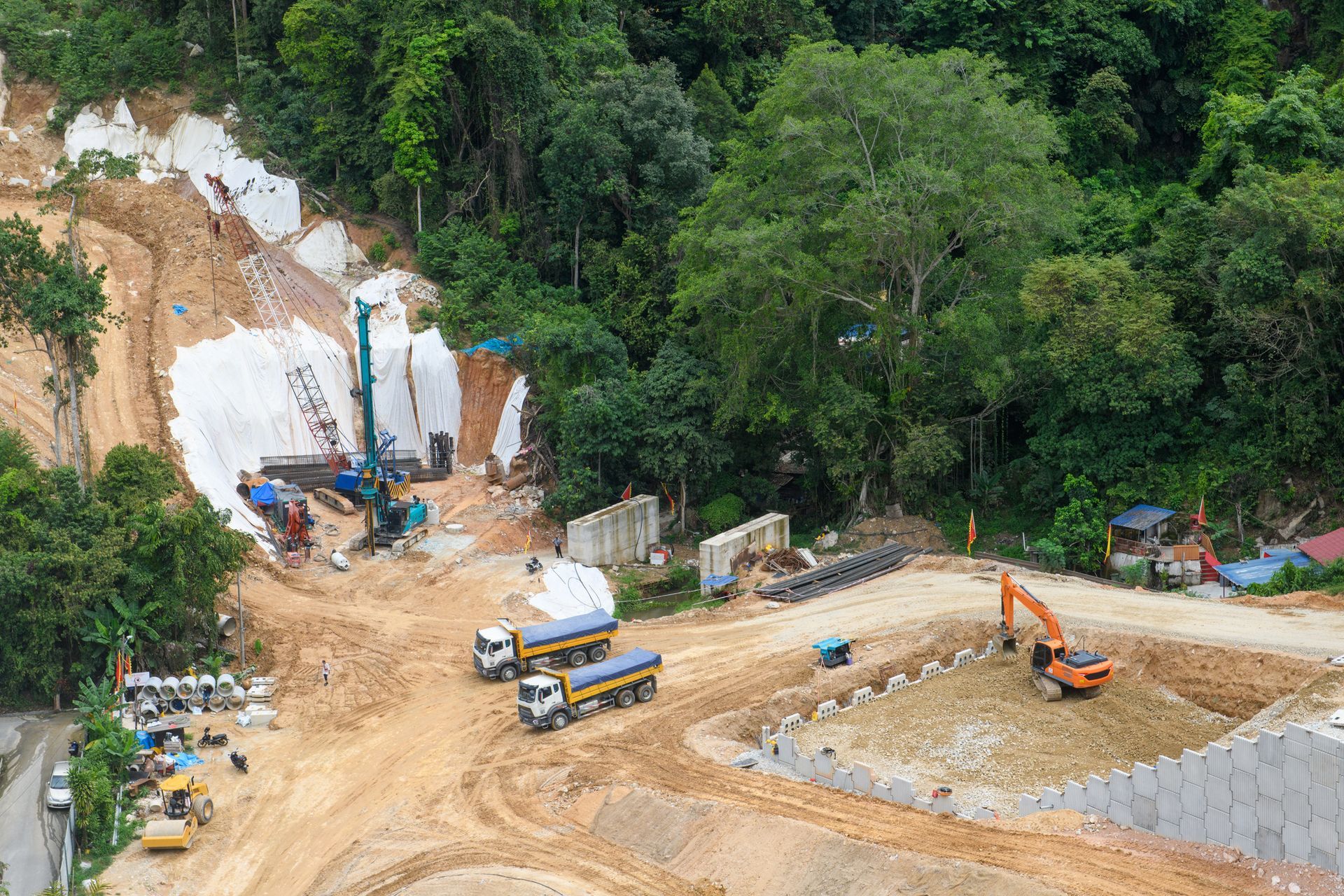 Construction site in a hillside, with machinery, trucks, and cut earth. Green trees surround the area.