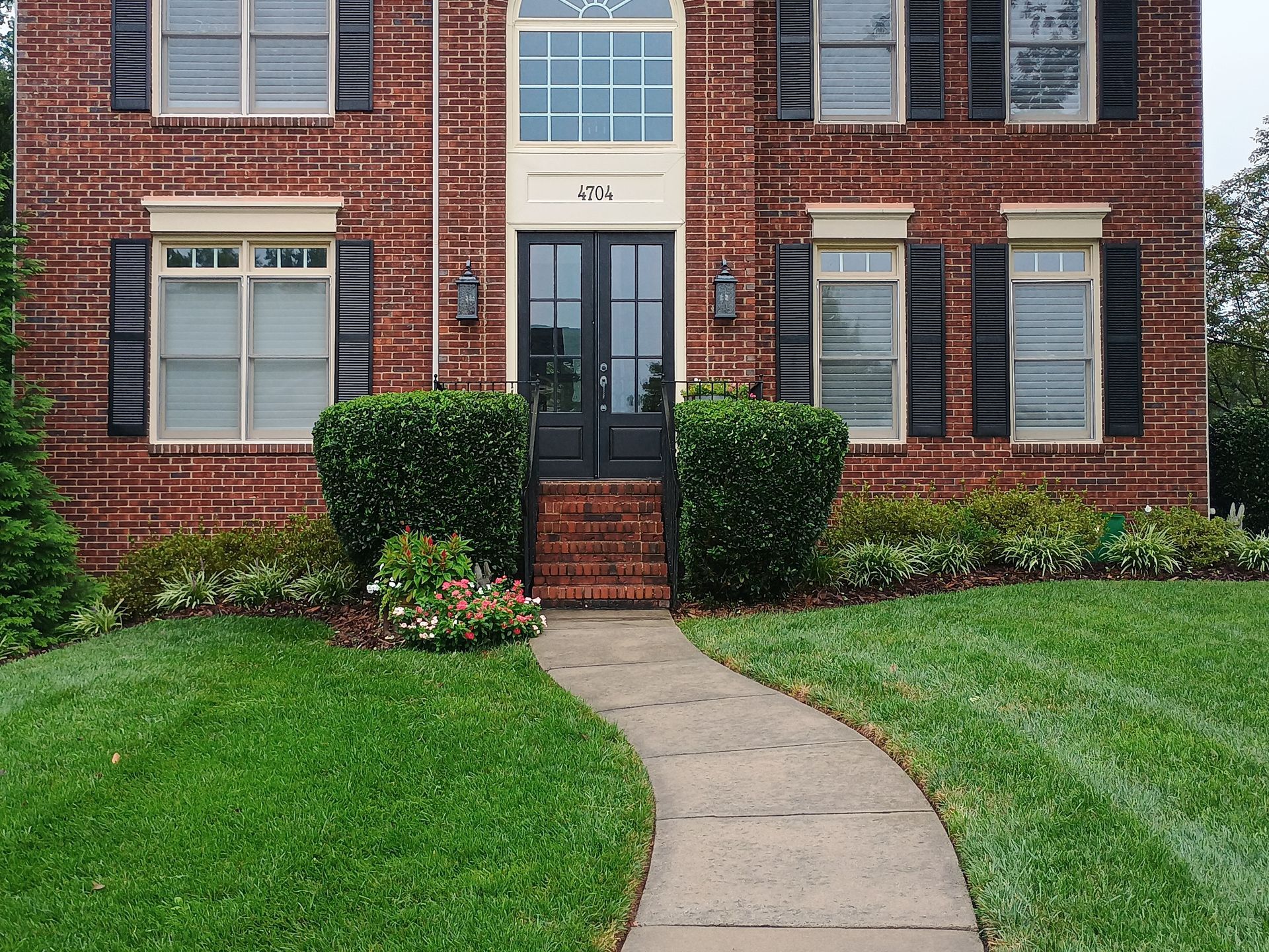 Brick house with black shutters and door; a curved path leads to the entrance, lush green lawn.