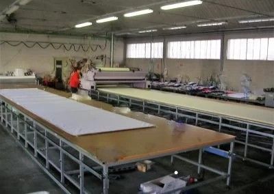 In uno stabilimento di produzione tessile, una persona con una giacca arancione lavora a un lungo tavolo, circondata da macchinari industriali.