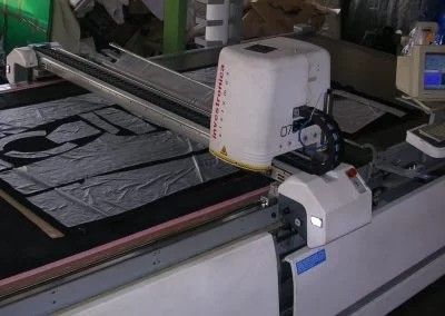 In una fabbrica, una macchina da taglio industriale con una testina bianca si muove su strati di tessuto stesi su un lungo tavolo.