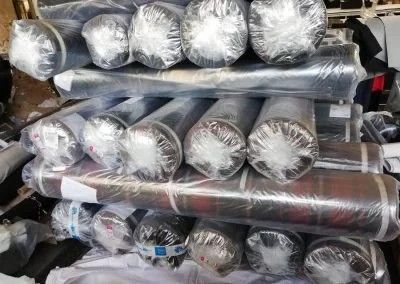 Rotoli di tessuto impilati e avvolti singolarmente in plastica trasparente, disposti in un magazzino.