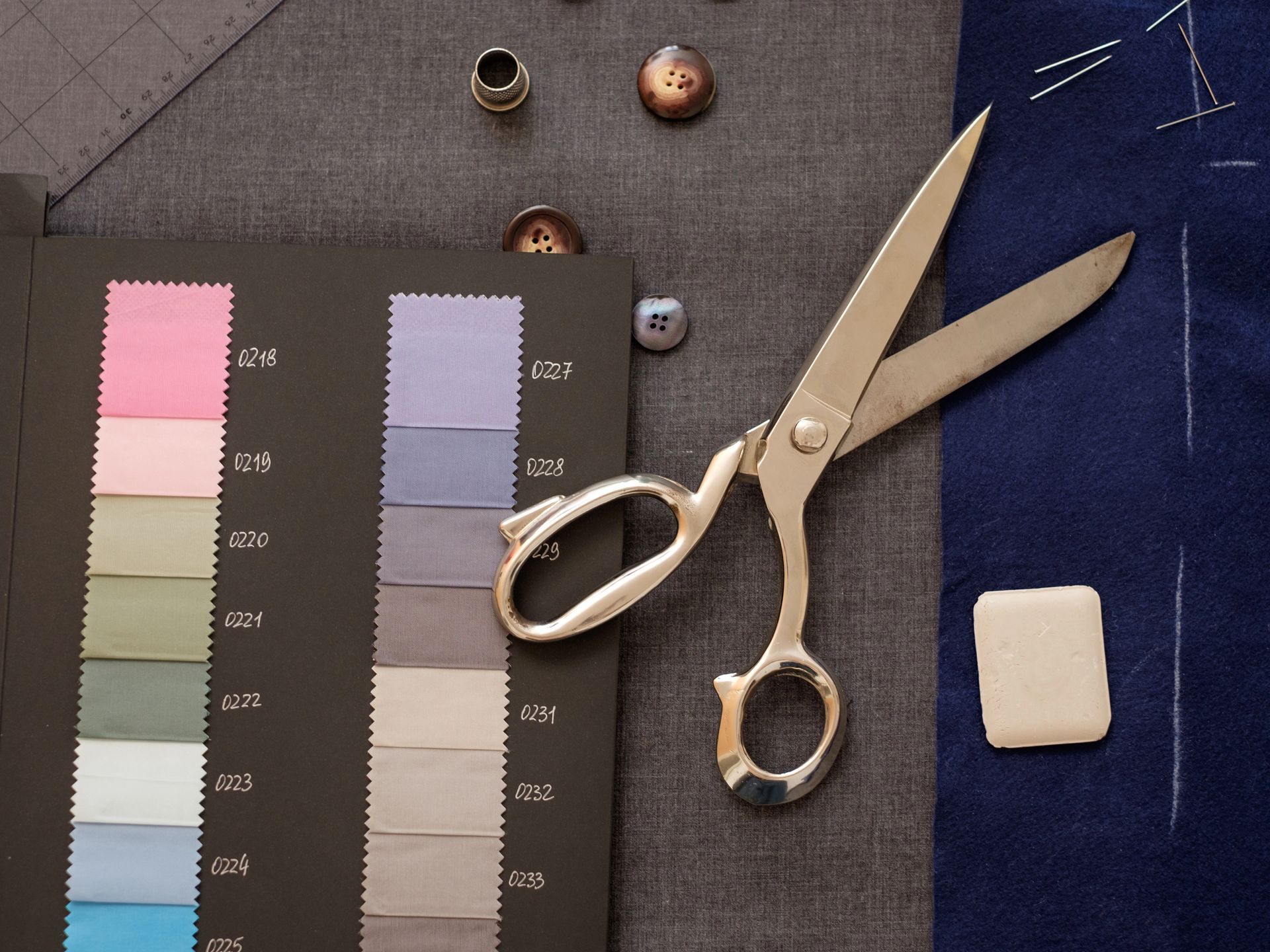 Un campionario di tessuti, delle forbici di metallo, dei bottoni e una squadra da sarto disposti su un tessuto grigio e blu.