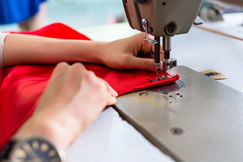 Delle mani guidano un tessuto rosso attraverso una macchina da cucire posta su un tavolo.