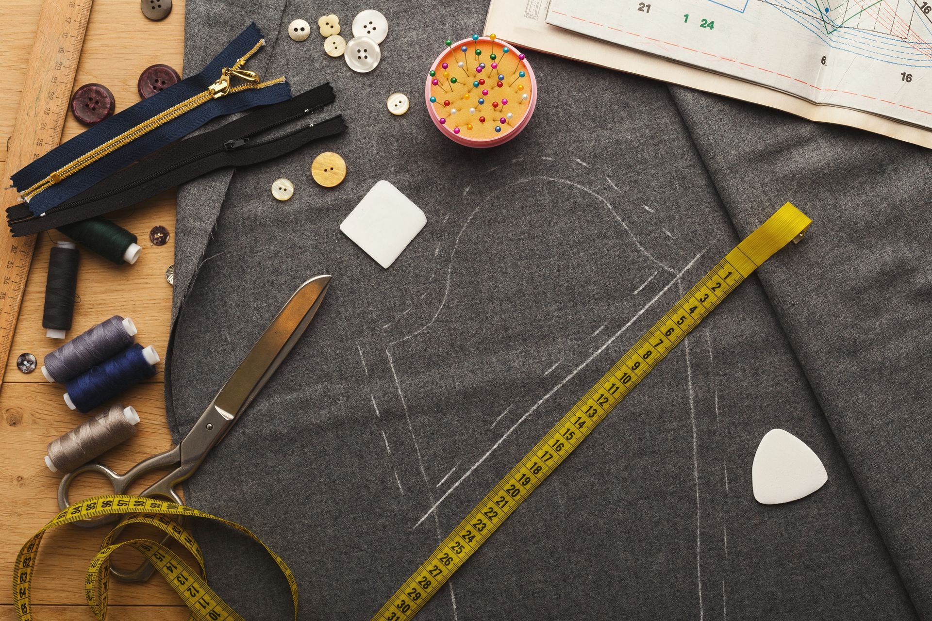 Materiali da sartoria, tra cui tessuto, cerniere, filo, metro a nastro, forbici e cartamodelli, disposti su un tavolo.