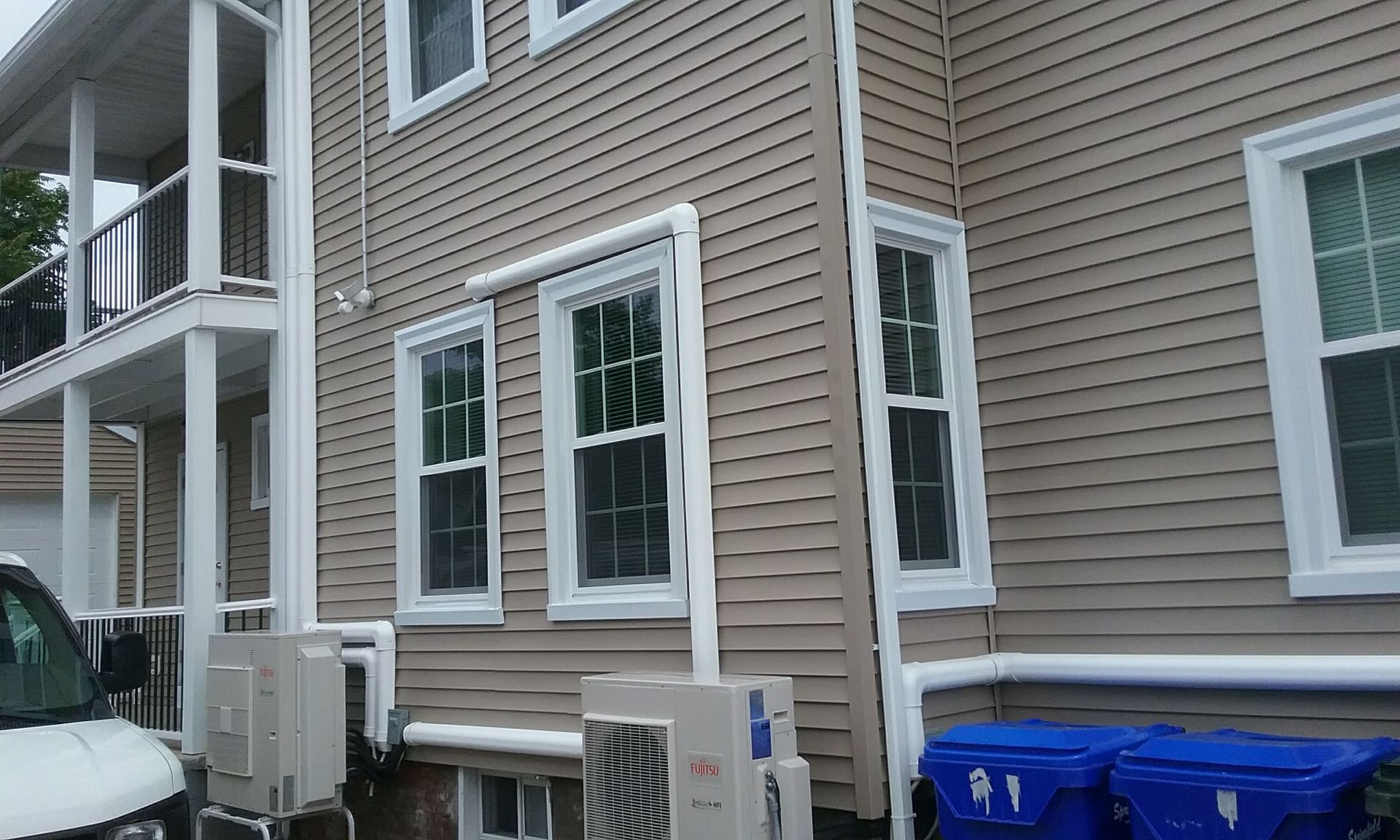 Tan house with white trim and windows. Air conditioning units and blue trash bins are at the base.