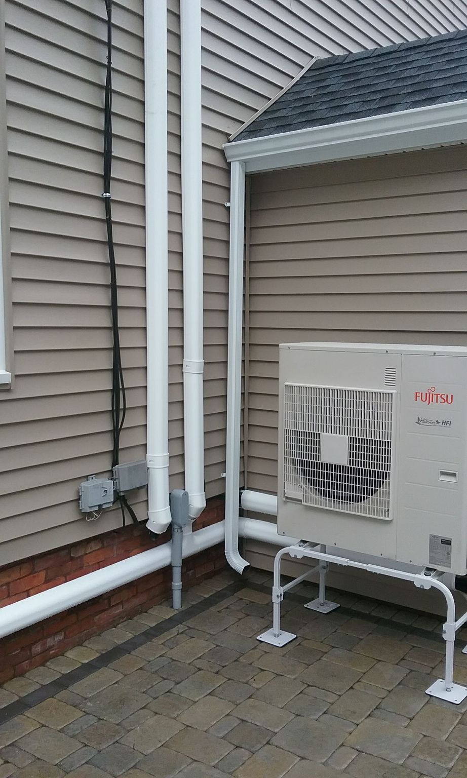 Exterior of a house with an air conditioning unit on a stand; white pipes and black electrical conduit on the wall.