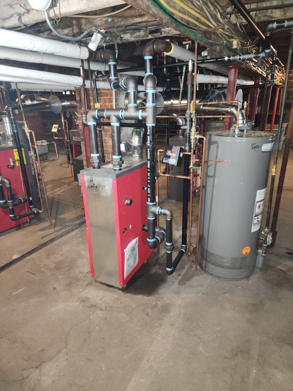 Red heating unit, gray water tank, and black pipes in a basement with a concrete floor.