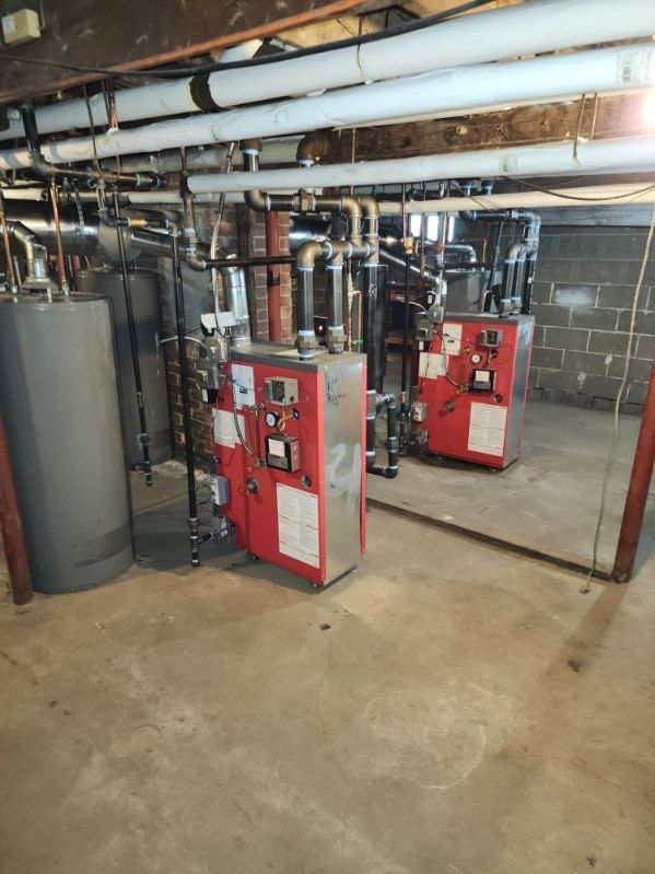 Basement mechanical room with two red furnaces, gray water heater, and pipes against a cement wall.