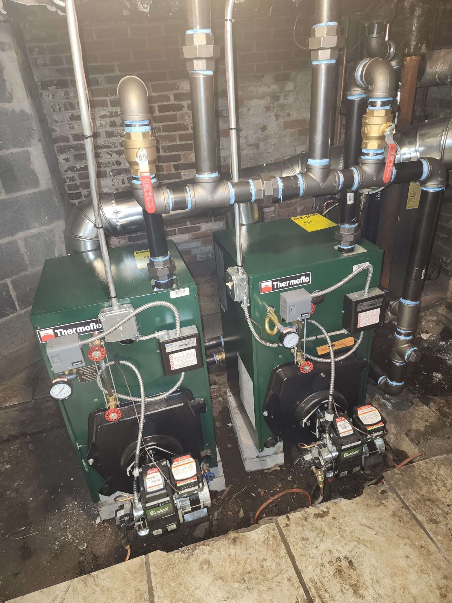 Two green industrial boilers with black components and silver piping in a brick-walled utility room.