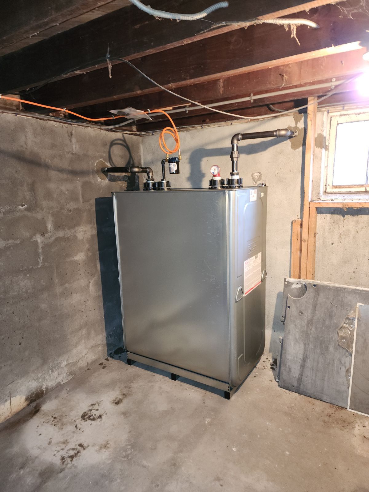 Gray furnace in a basement, with pipes and wires against a concrete wall and wooden beams.