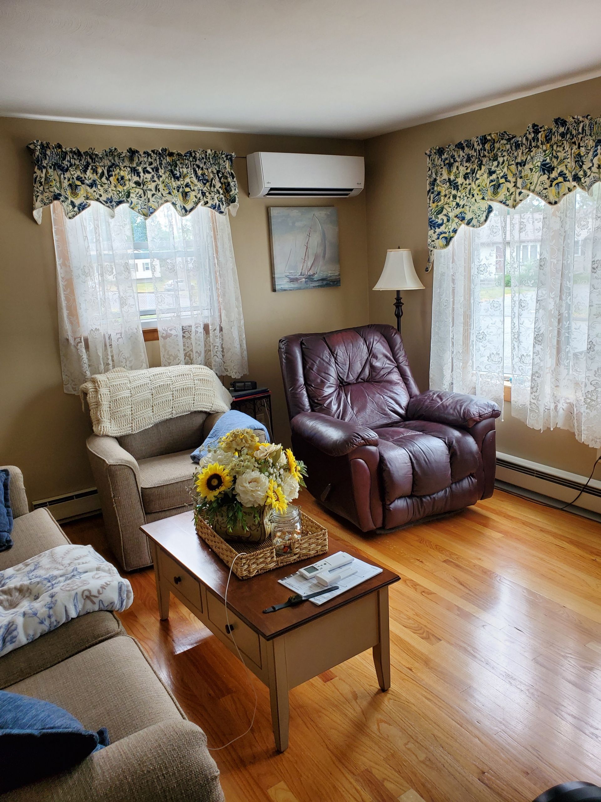 Living room with hardwood floors, two windows with lace curtains, a leather recliner, and a beige sofa.