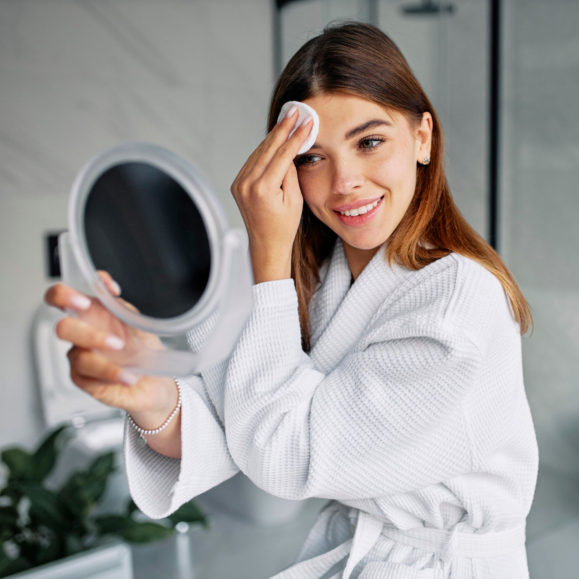 A woman in a bathrobe is cleaning her face in front of a mirror.