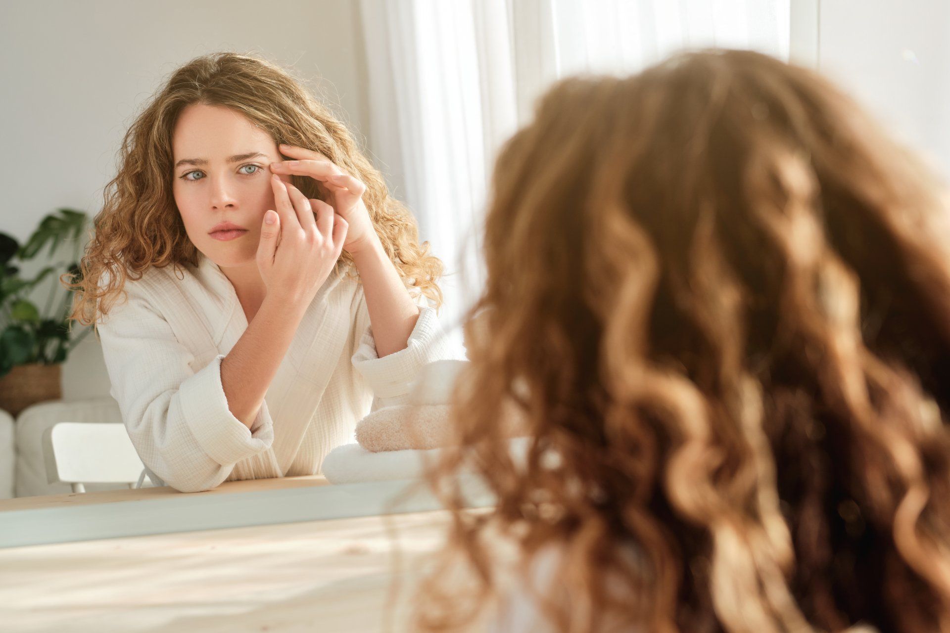 woman with curly hair examining eye wrinkles in mirror
