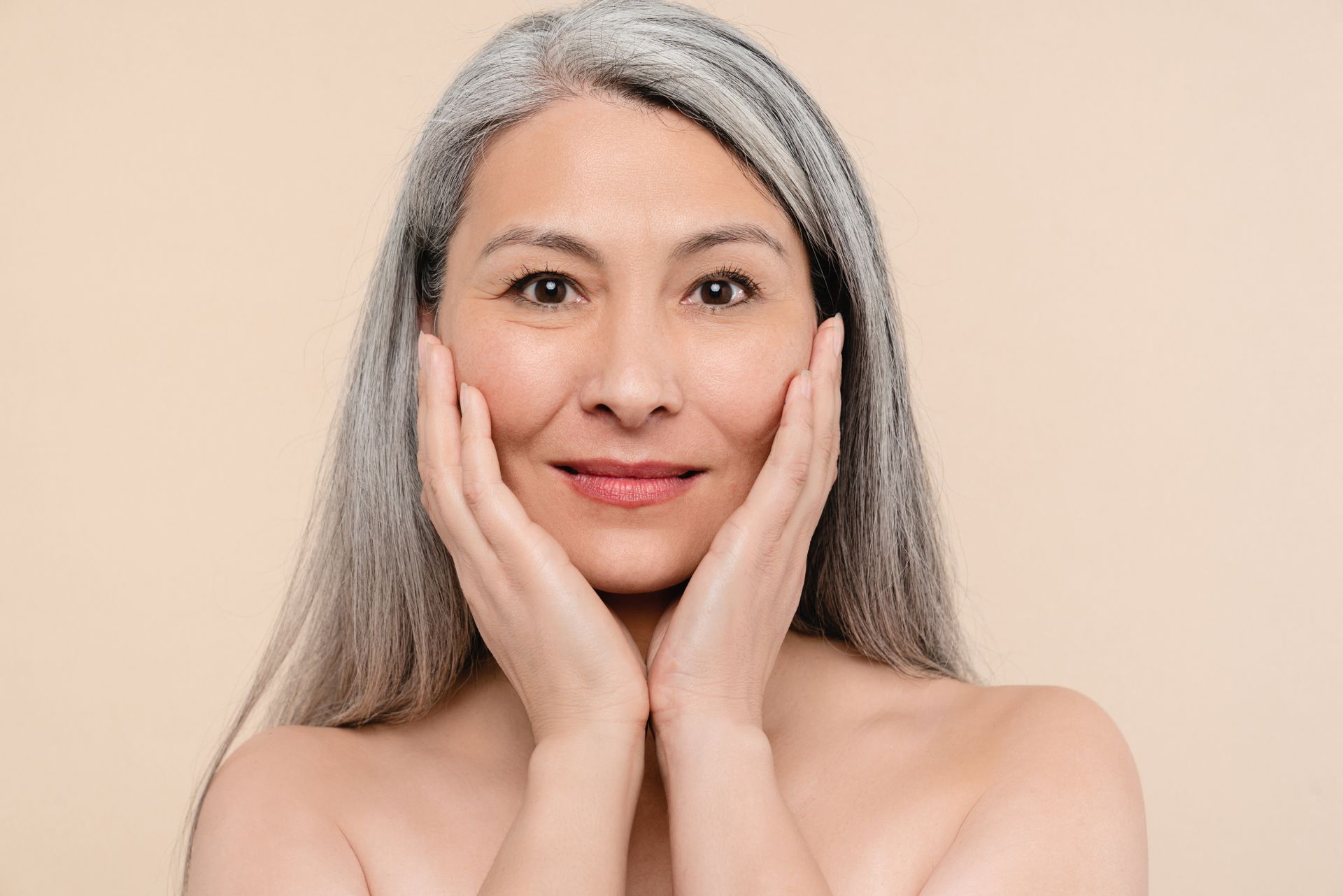 A woman with gray hair is smiling and touching her face with her hands.