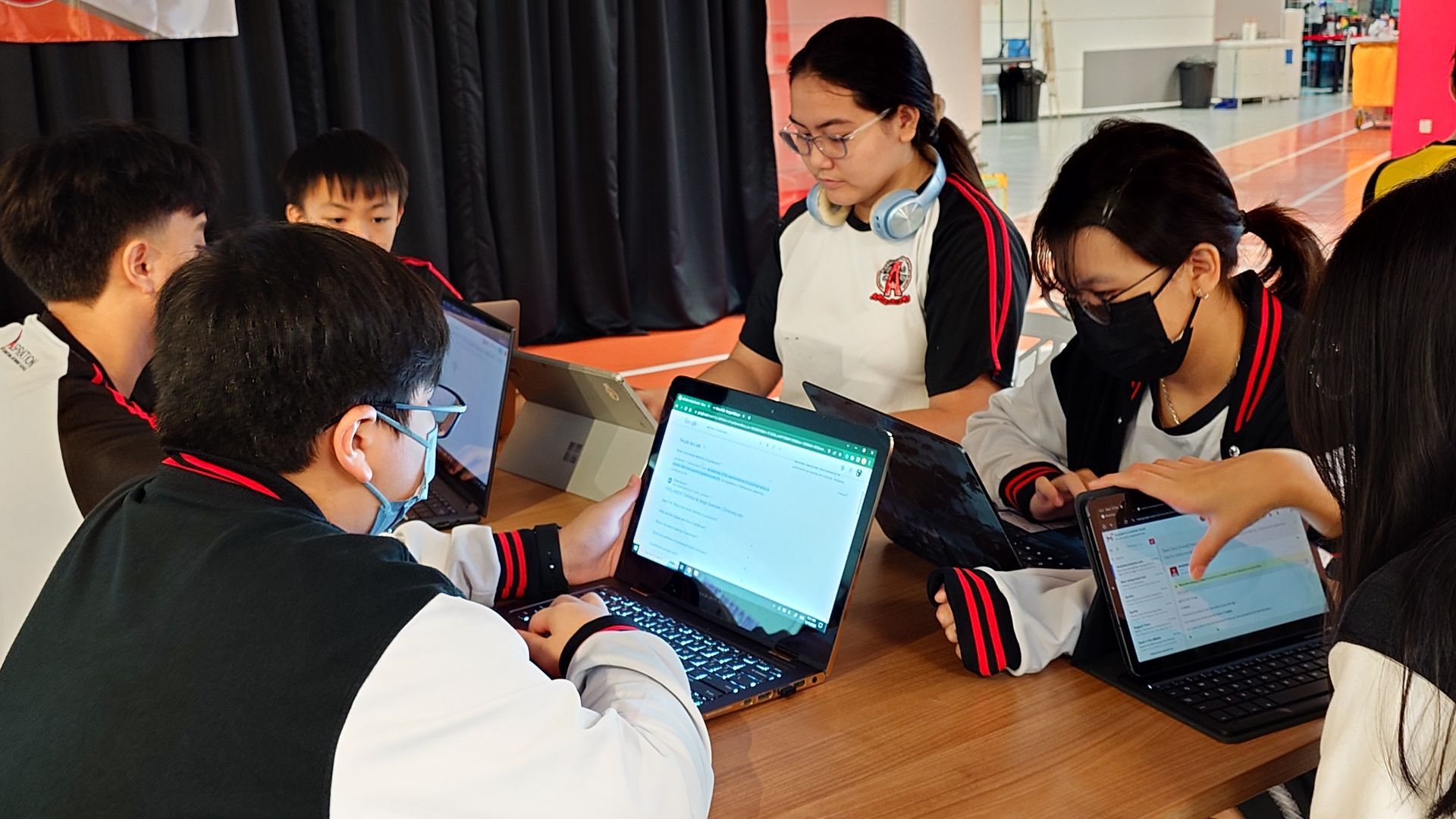 Students in school uniforms work on laptops at a table, some wearing masks.