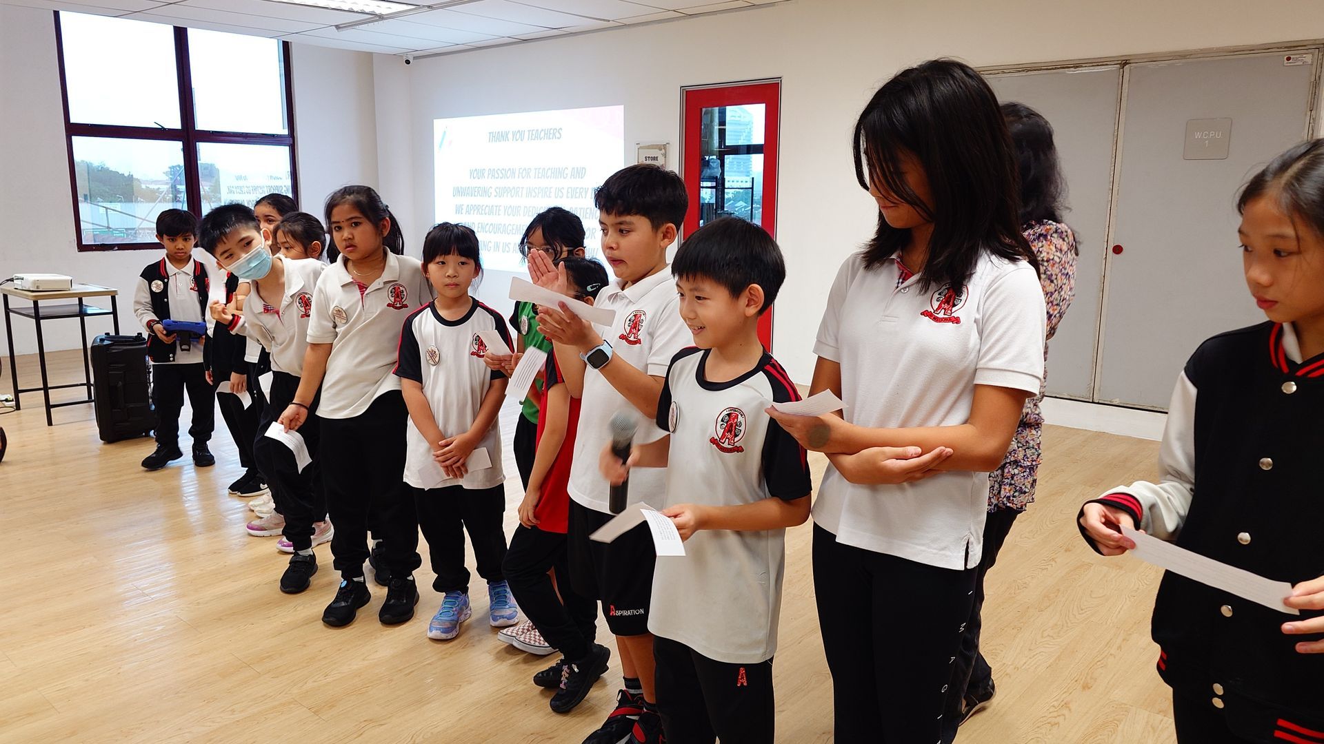 Children standing in a classroom, some holding papers, and one reading aloud. A screen is in the background.