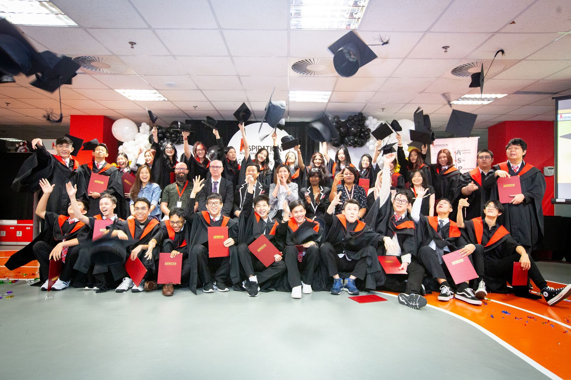 Graduation celebration: group in caps and gowns toss hats in the air. Red and black accents, holding diplomas.