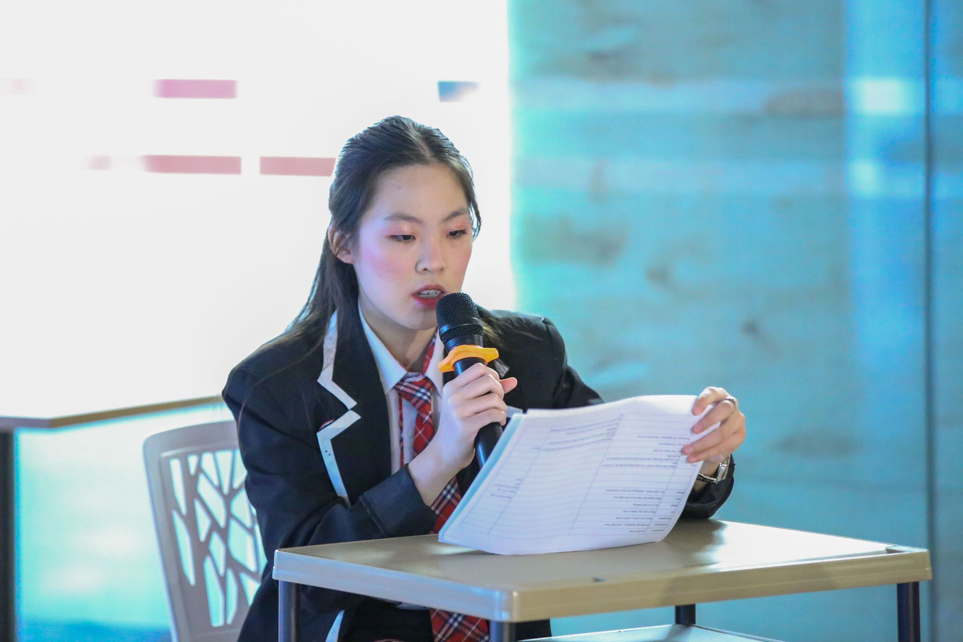 Young person in blazer speaking into a microphone while reading from papers at a desk.