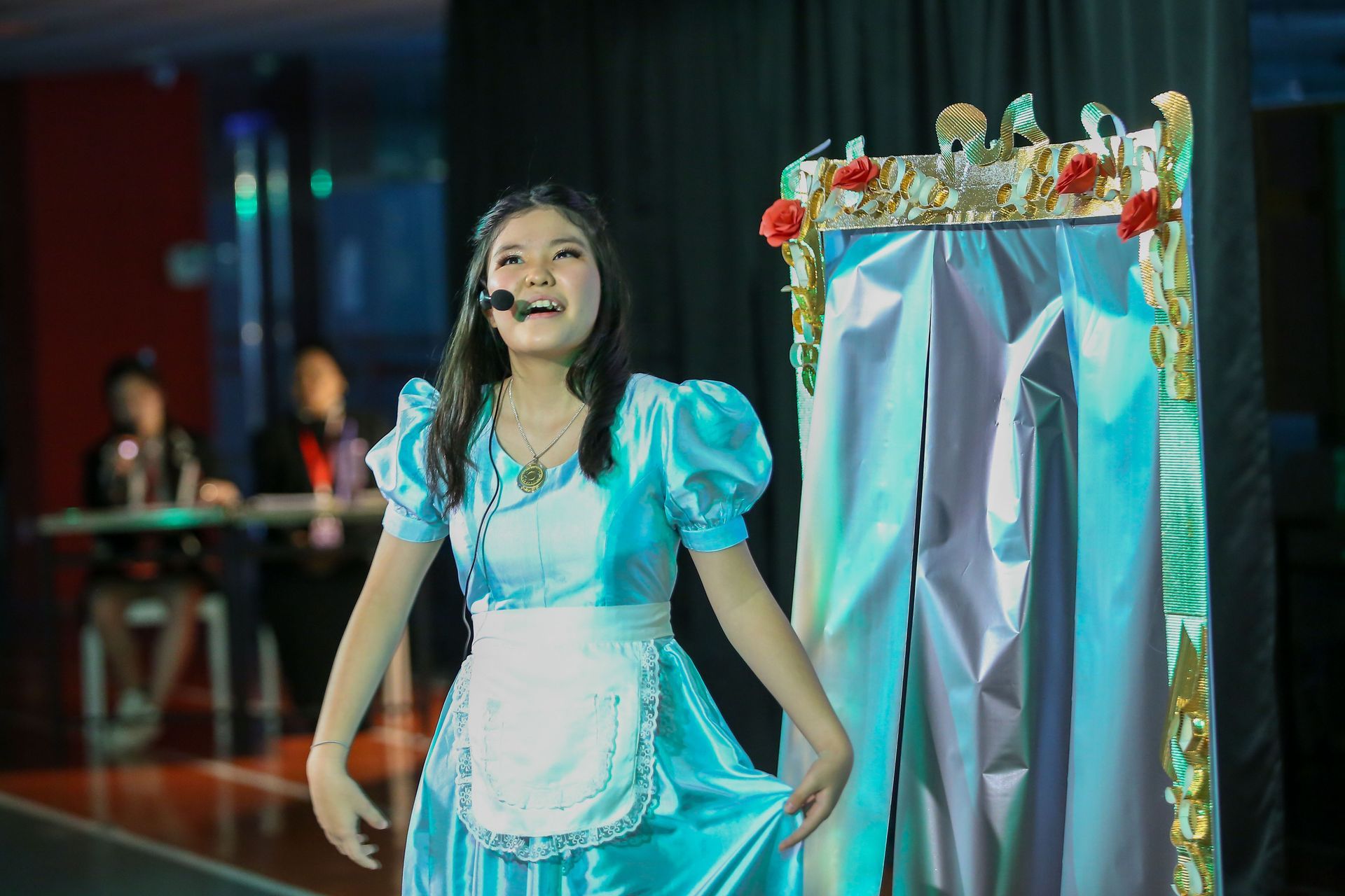 A person in a blue dress and apron, singing with a stage backdrop, smiling.
