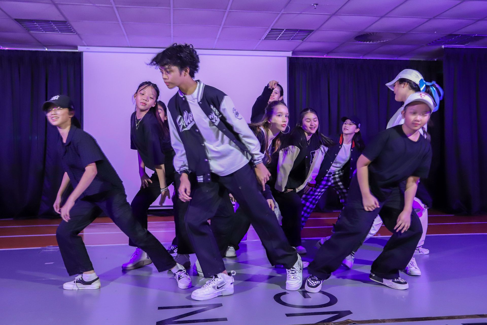 Dancers in a studio, performing a hip-hop routine. They wear black and white, lit by purple lights.