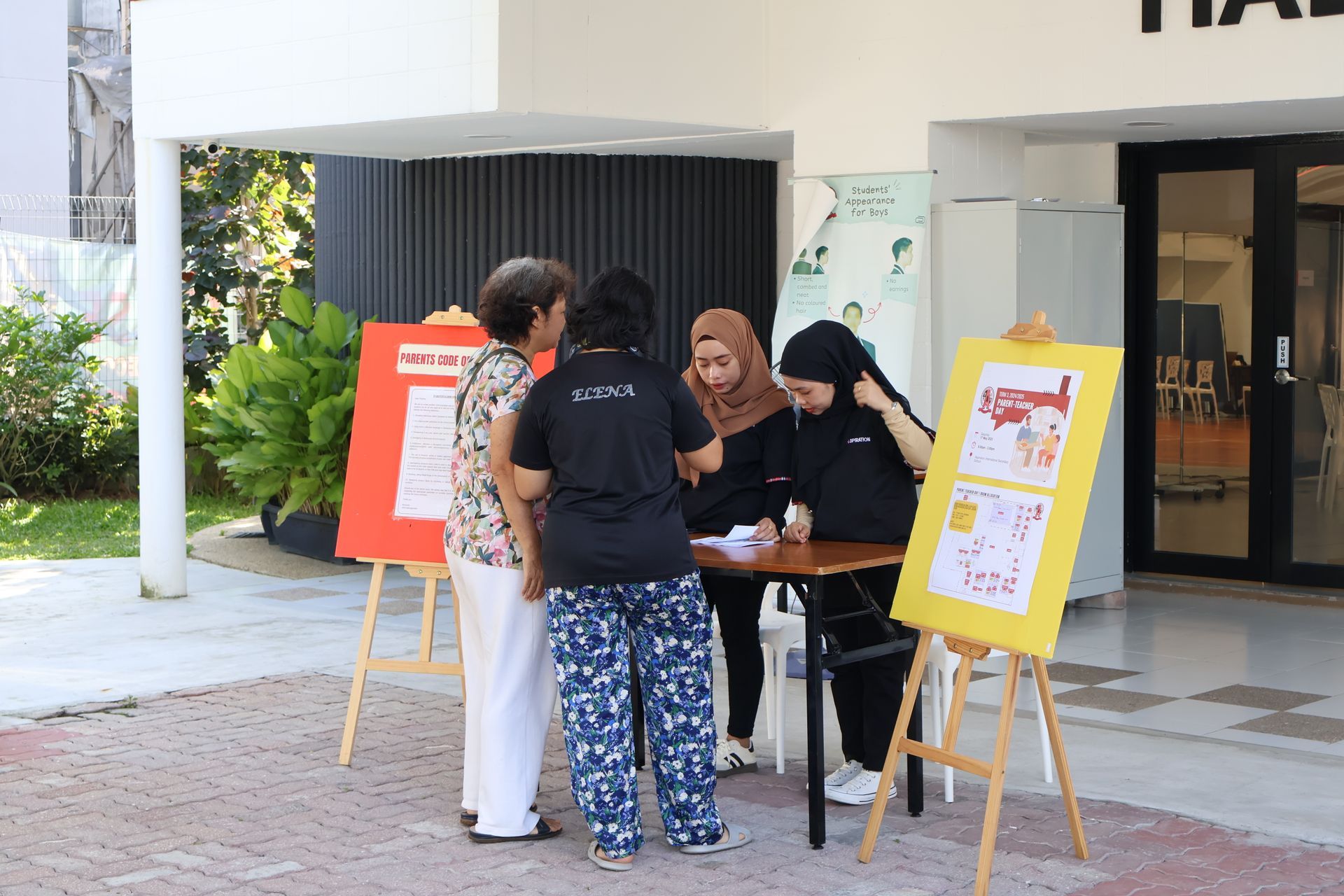 People examining posters at an outdoor event, some wearing black shirts and headscarves, near a building.