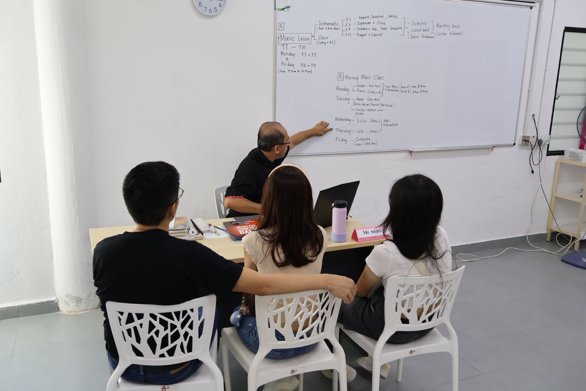 Teacher pointing at whiteboard, explaining math to three students seated at a table. White room.