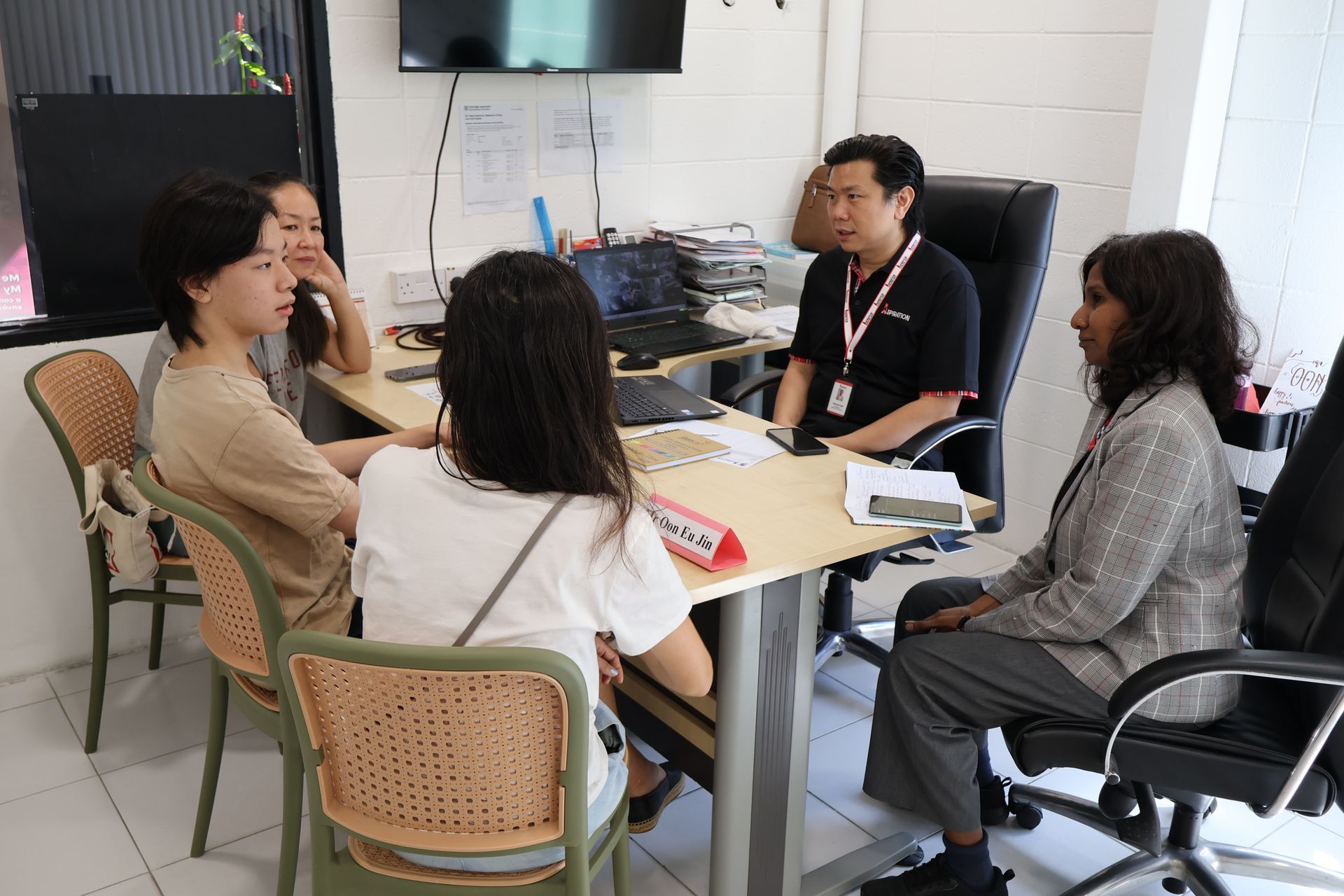 People seated at a table, discussing. Man in red shirt, woman in gray suit. Room with computer, monitor.