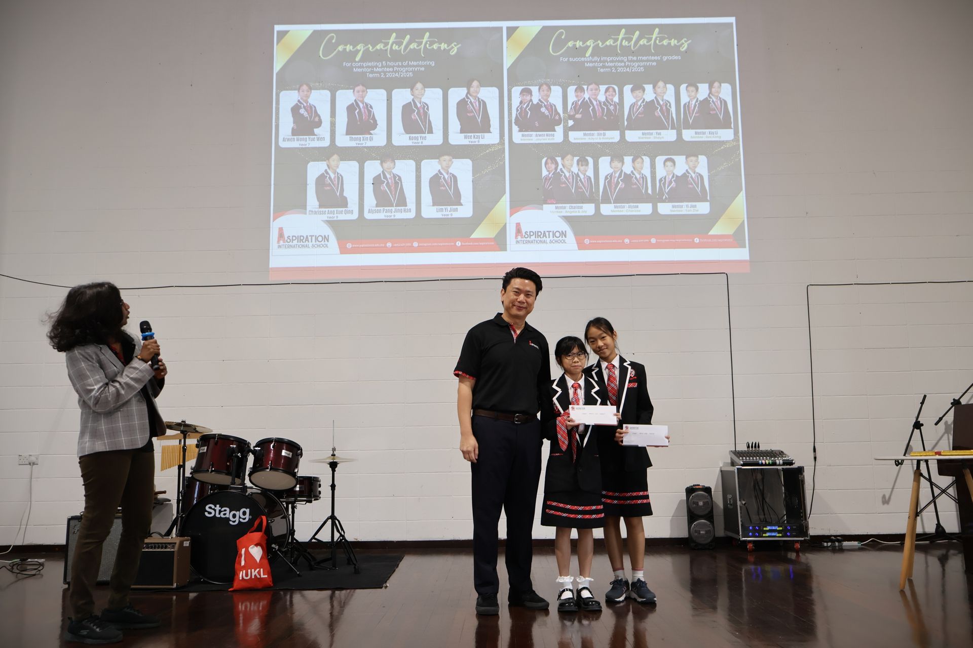 A presentation onstage with a man presenting certificates to two schoolgirls, with a projector screen showing photos.